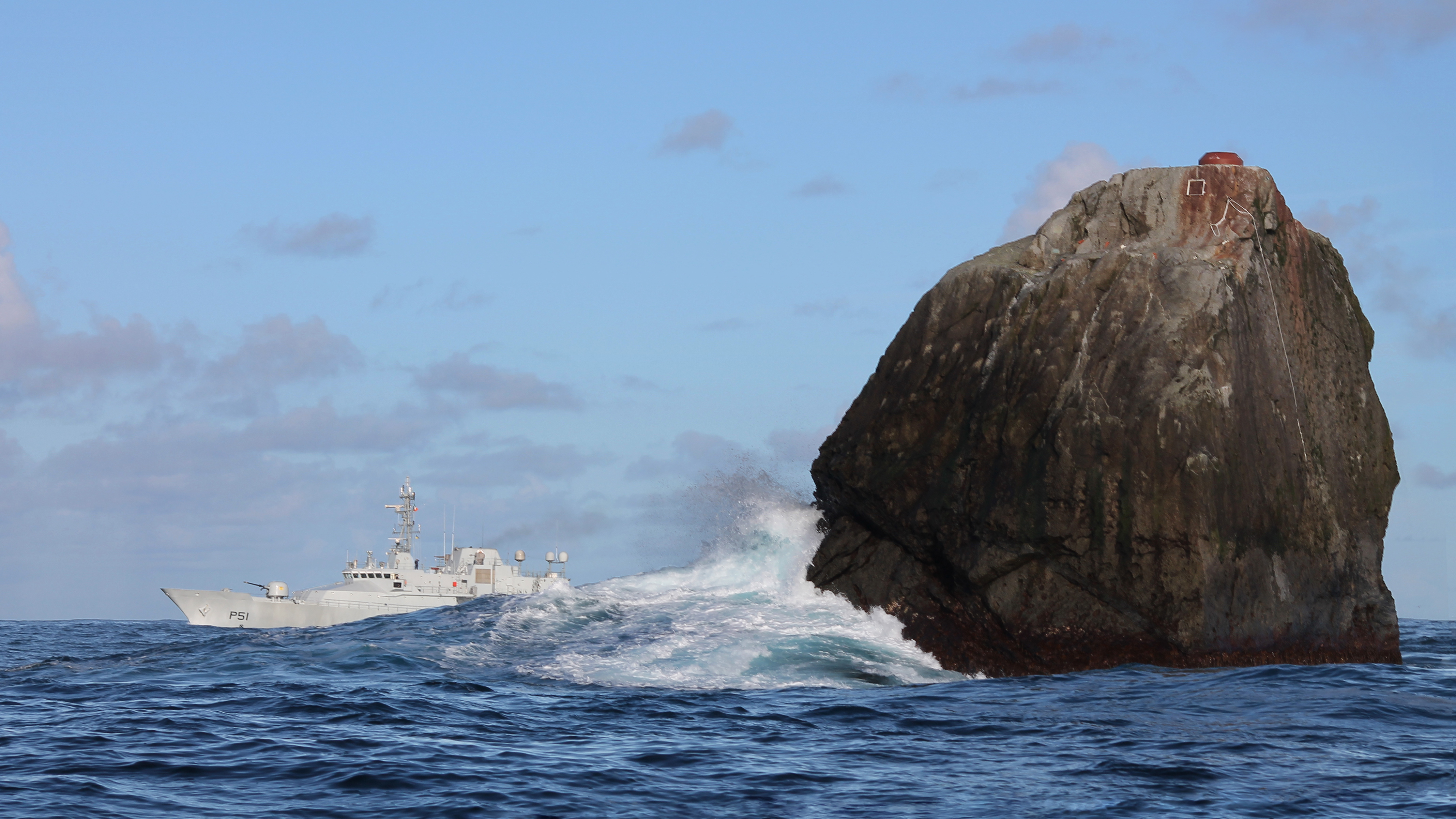 Atlantic waves breaking against Rockall, with the patrol vessel LÉ Róisín in the distance
